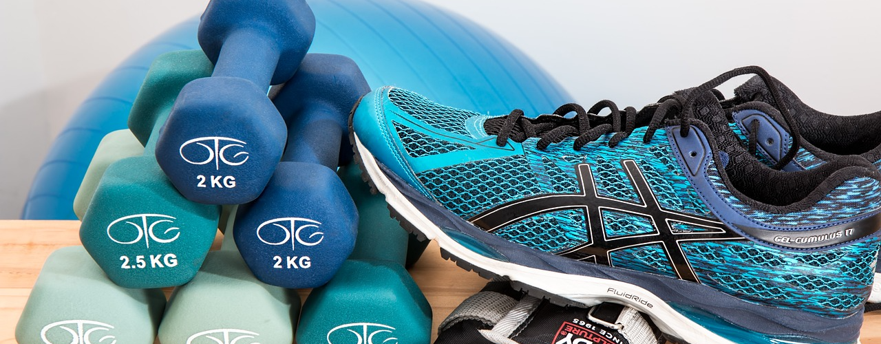 Eine Sammlung von Fitnessutensilien auf einem Holztisch: blaue und grüne Hanteln, ein Paar blaue Sportschuhe, Fitnesshandschuhe und ein großer Gymnastikball im Hintergrund.