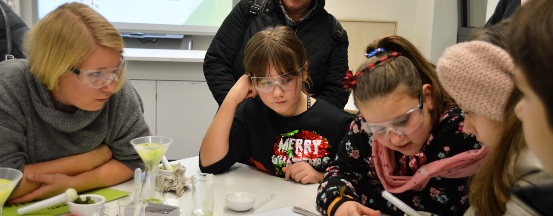 Eine Gruppe von Kindern und Erwachsenen, ausgestattet mit Schutzbrillen, sitzt an einem Tisch und führt ein wissenschaftliches Experiment durch. Im Hintergrund ist eine Präsentation über Pflanzen und Zellen zu sehen