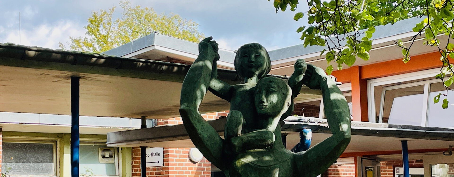 Eine Statue zeigt eine Person, die ein Kind auf den Schultern trägt, vor einem Backsteingebäude mit einem Flachdach und Bäumen im Vordergrund