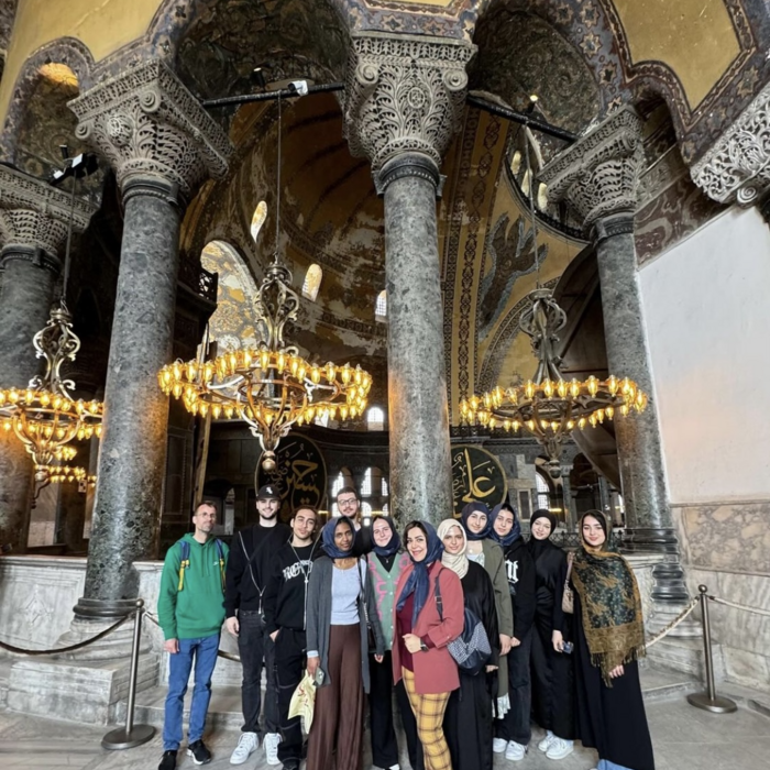 Eine Gruppe von Menschen posiert in einem historischen Gebäude mit Säulen und Kronleuchtern. Der Raum hat ein goldenes Dach und ein kunstvolles Design