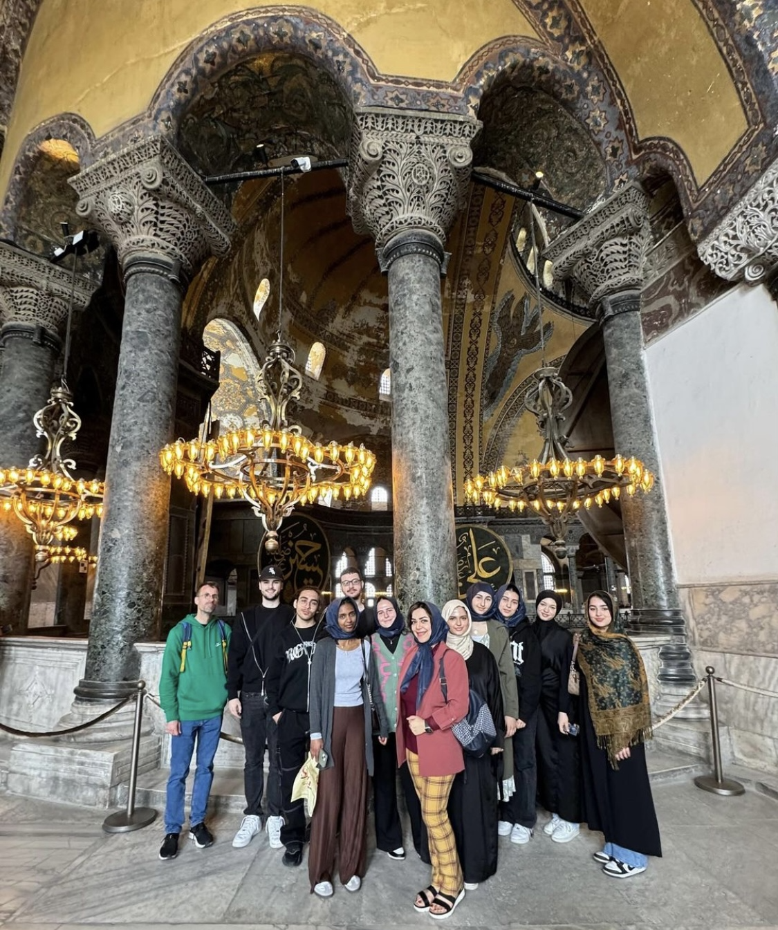 Eine Gruppe von Menschen posiert in einem historischen Gebäude mit Säulen und Kronleuchtern. Der Raum hat ein goldenes Dach und ein kunstvolles Design