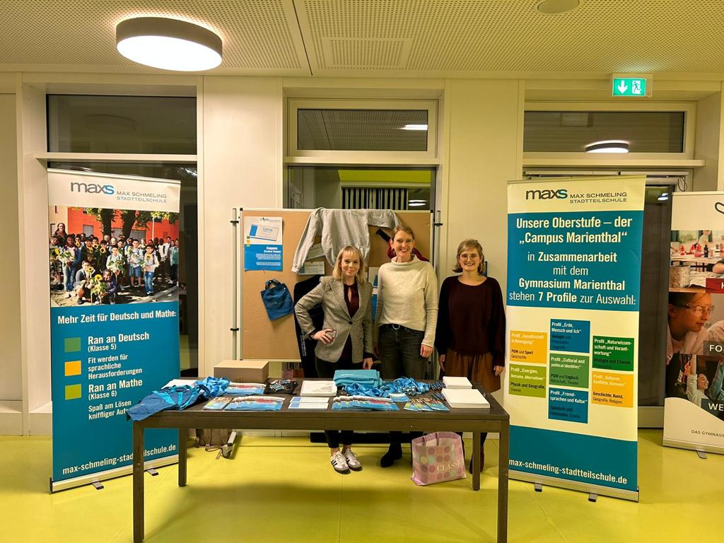 Drei Frauen stehen an einem Informationsstand der Max-Schmeling-Stadtteilschule, flankiert von Roll-Ups mit Informationen zu Schulprogrammen. Auf dem Tisch liegen Flyer und kleine Geschenke aus