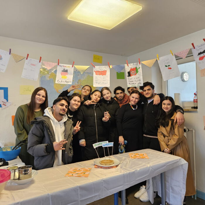 Eine Gruppe junger Menschen posiert fröhlich hinter einem dekorierten Tisch mit einem Kuchen. Der Raum ist mit Wimpeln und Postern geschmückt, die verschiedene Länder und Kulturen repräsentieren.