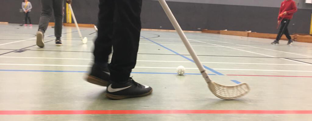 Schüler spielen in einer Turnhalle Floorball. Im Vordergrund ist ein Spieler mit Schläger und Ball zu sehen, während weitere Spieler im Hintergrund aktiv sind.