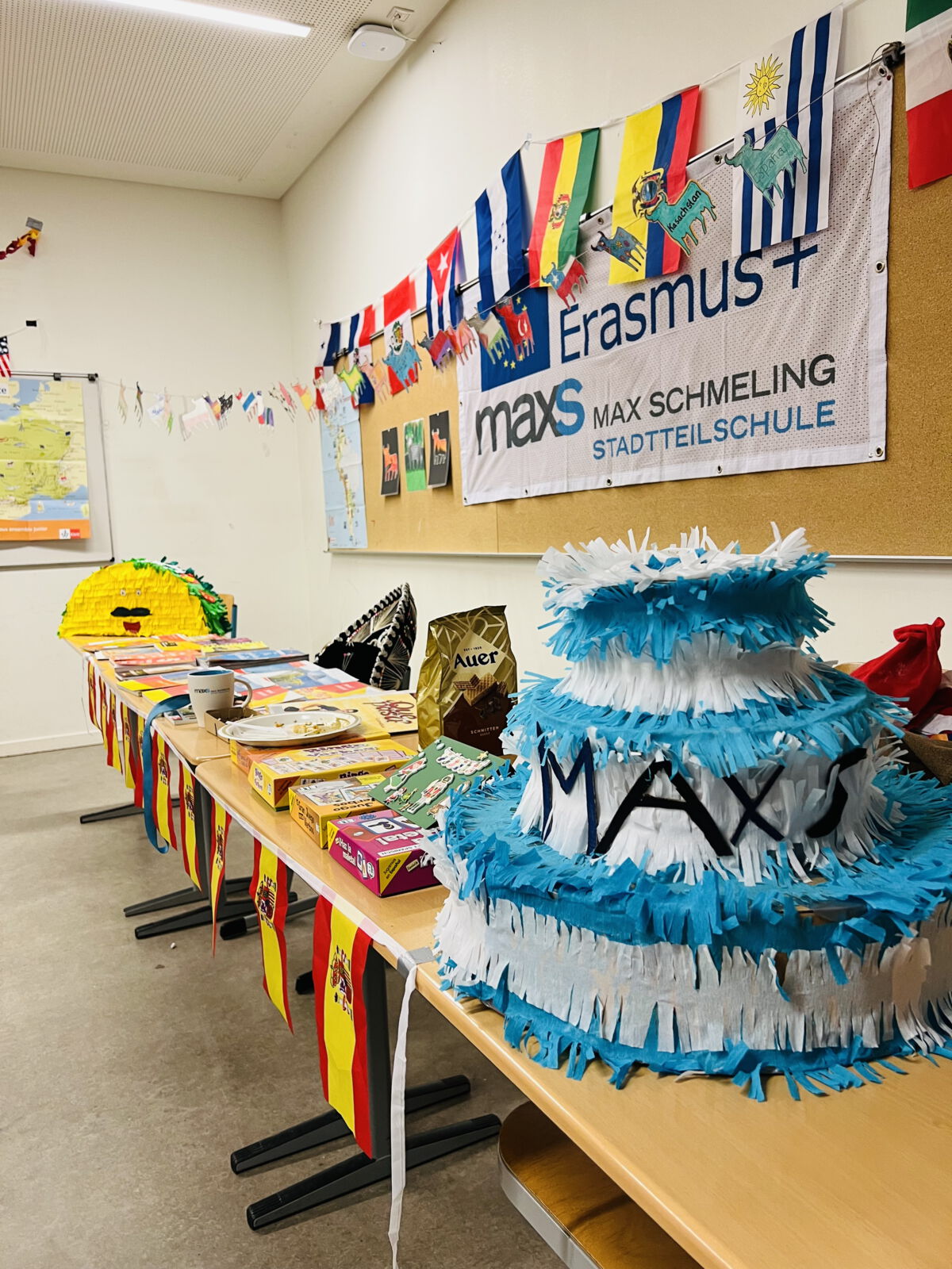 Ein Raum mit Erasmus+-Banner der Max-Schmeling-Stadtteilschule. Auf einem Tisch stehen Spiele, Snacks und eine blau-weiße Piñata mit 'MAX'. Flaggen und Zeichnungen dekorieren den Raum