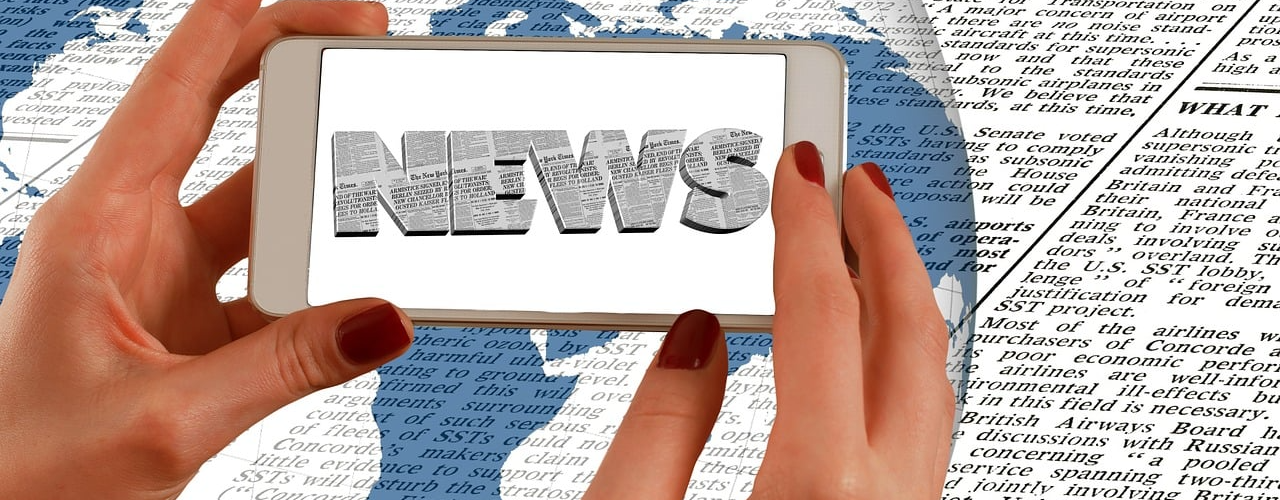 Zwei Hände halten ein Smartphone mit dem Schriftzug 'NEWS' auf dem Bildschirm. Im Hintergrund sind eine Weltkarte und Zeitungsartikel zu sehen