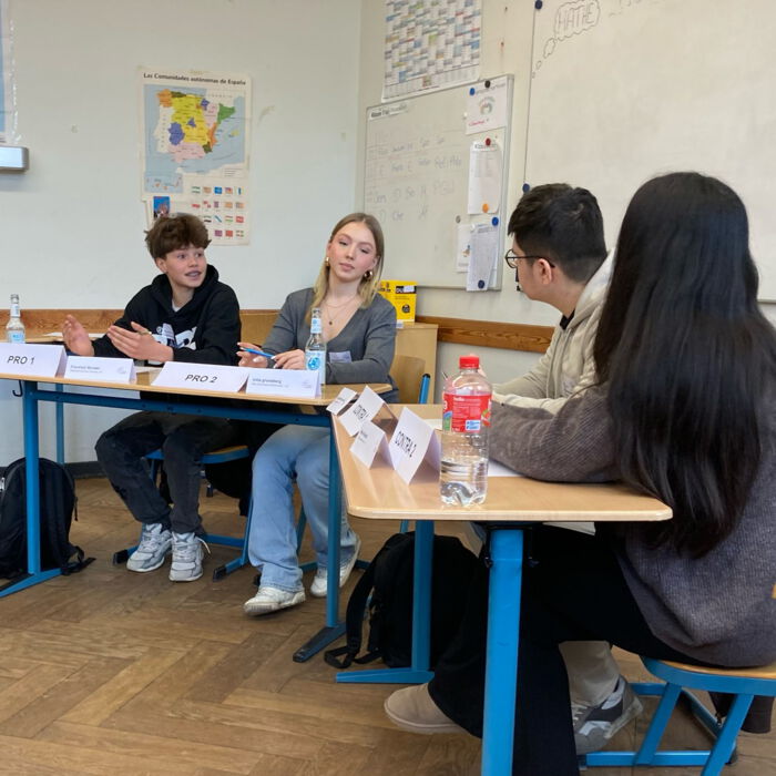 Vier Schüler*innen sitzen an Tischen in einem Klassenzimmer und diskutieren engagiert. Vor ihnen stehen Namensschilder