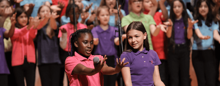 Ein Kinderchor mit bunten T-Shirts singt auf einer Bühne, während zwei Mädchen vorne mit Mikrofonen performen