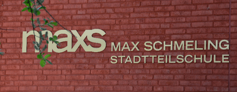 Logo der Max-Schmeling-Stadtteilschule auf einer roten Ziegelwand. Der Schriftzug 'maxs MAX SCHMELING STADTTEILSCHULE' ist in goldener Schrift zu sehen, mit Zweigen im Vordergrund.