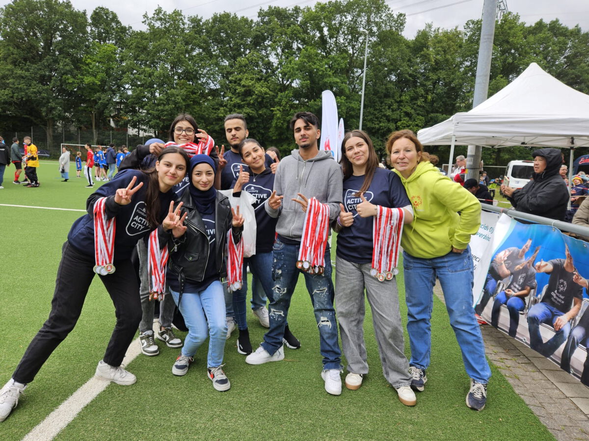 Eine Gruppe junger Menschen posiert fröhlich auf einem Sportplatz mit Medaillen. Sie tragen T-Shirts mit der Aufschrift 'Active City' und zeigen Peace-Zeichen. Im Hintergrund sind ein Fußballspiel und Zuschauer zu sehen