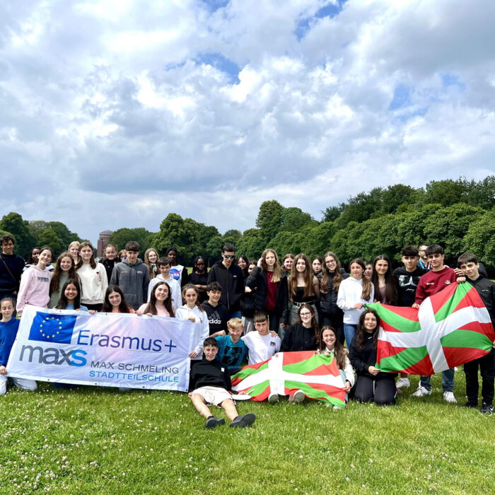 Eine große Gruppe von Schüler*innen steht auf einer Wiese unter einem bewölkten Himmel. Sie halten ein Erasmus+-Banner der Max-Schmeling-Stadtteilschule sowie baskische Flaggen