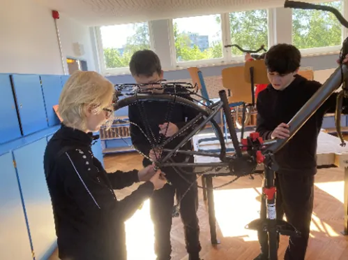 Drei Schüler arbeiten an einem Fahrrad, während es auf einem Ständer in einem Klassenraum befestigt ist