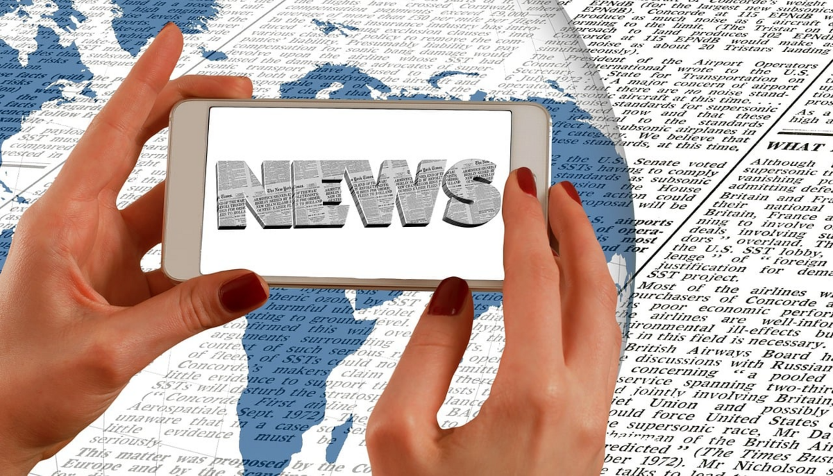 Zwei Hände halten ein Smartphone mit dem Schriftzug 'NEWS' auf dem Bildschirm. Im Hintergrund sind eine Weltkarte und Zeitungsartikel zu sehen