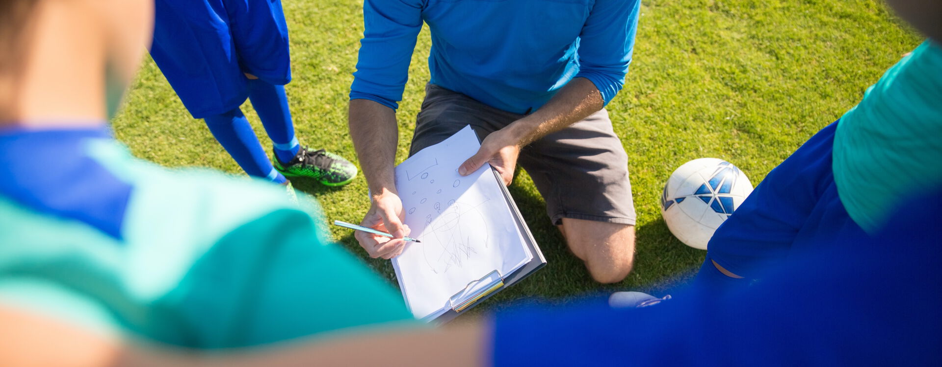 Ein Trainer kniet auf einem Fußballfeld und erklärt seinem Team etwas auf einem Klemmbrett. Um ihn stehen Kinder in Sportkleidung, ein Ball liegt daneben.