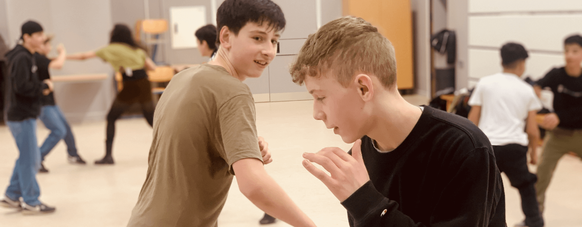 Zwei Jungen nehmen an einer Bewegungseinheit oder einem Tanzworkshop teil, während weitere Jugendliche im Hintergrund aktiv sind