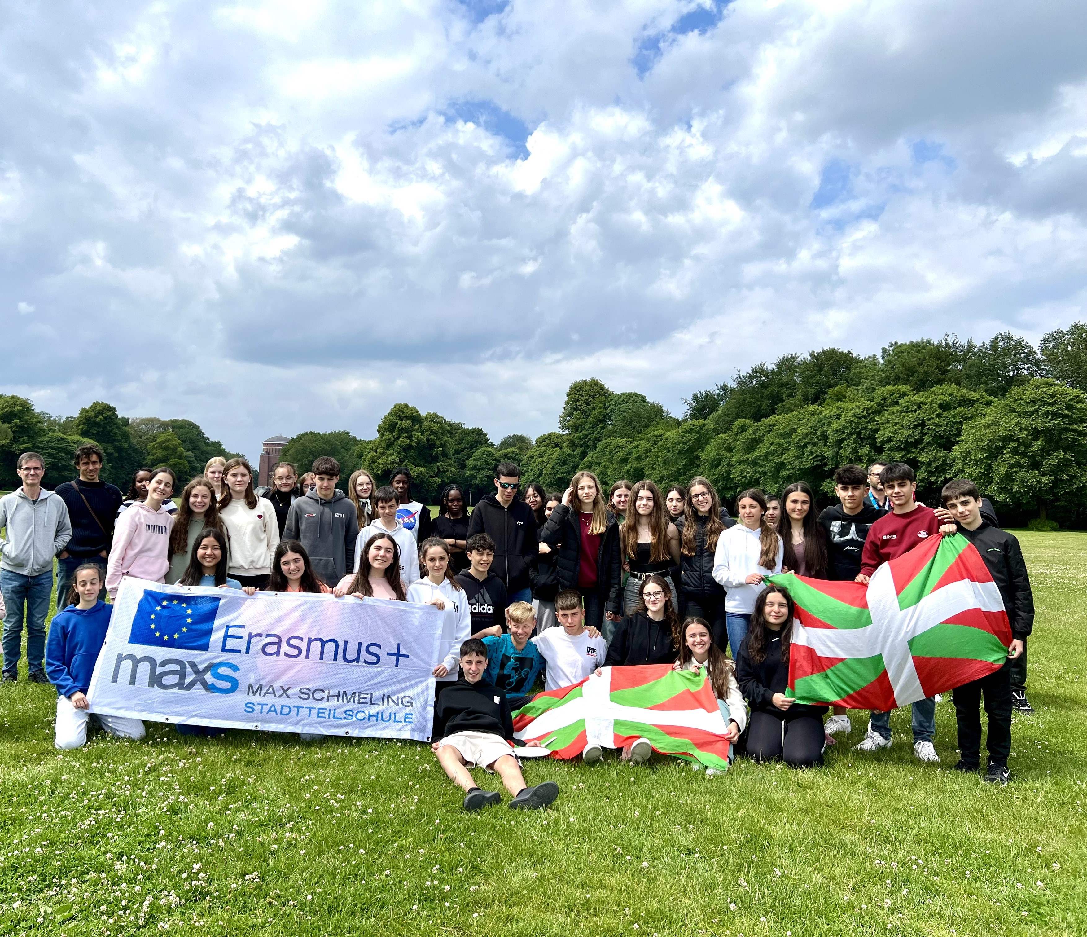 Eine große Gruppe von Schüler*innen steht auf einer Wiese unter einem bewölkten Himmel. Sie halten ein Erasmus+-Banner der Max-Schmeling-Stadtteilschule sowie baskische Flaggen