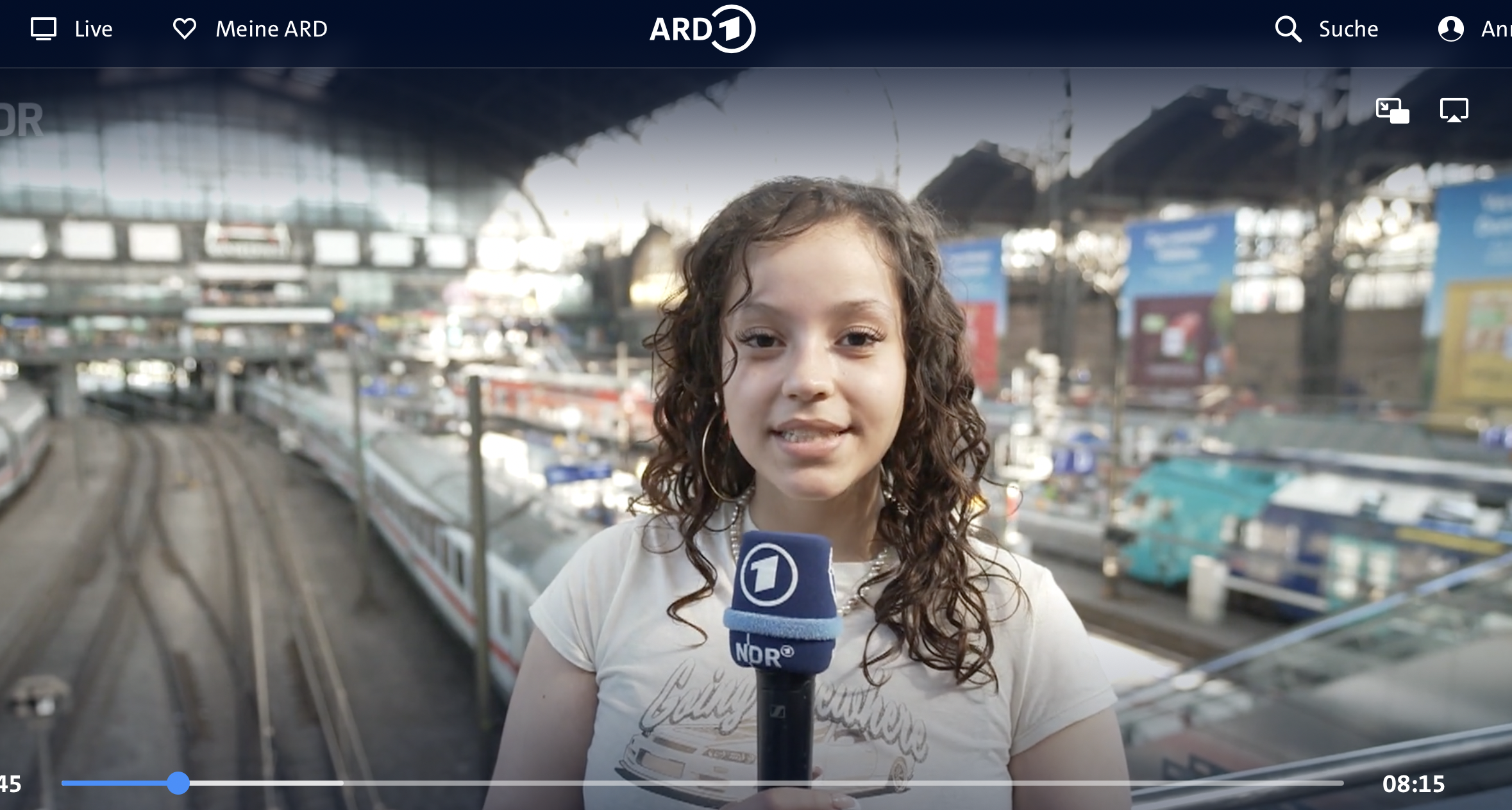 Ein junges Mädchen hält ein Mikrofon mit dem NDR-Logo und spricht in die Kamera. Im Hintergrund ist ein belebter Bahnhof mit Zügen und Gleisen zu sehen.
