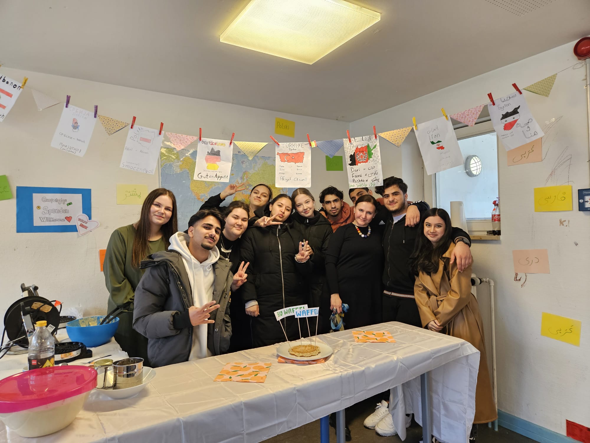 Eine Gruppe junger Menschen posiert fröhlich hinter einem dekorierten Tisch mit einem Kuchen. Der Raum ist mit Wimpeln und Postern geschmückt, die verschiedene Länder und Kulturen repräsentieren.
