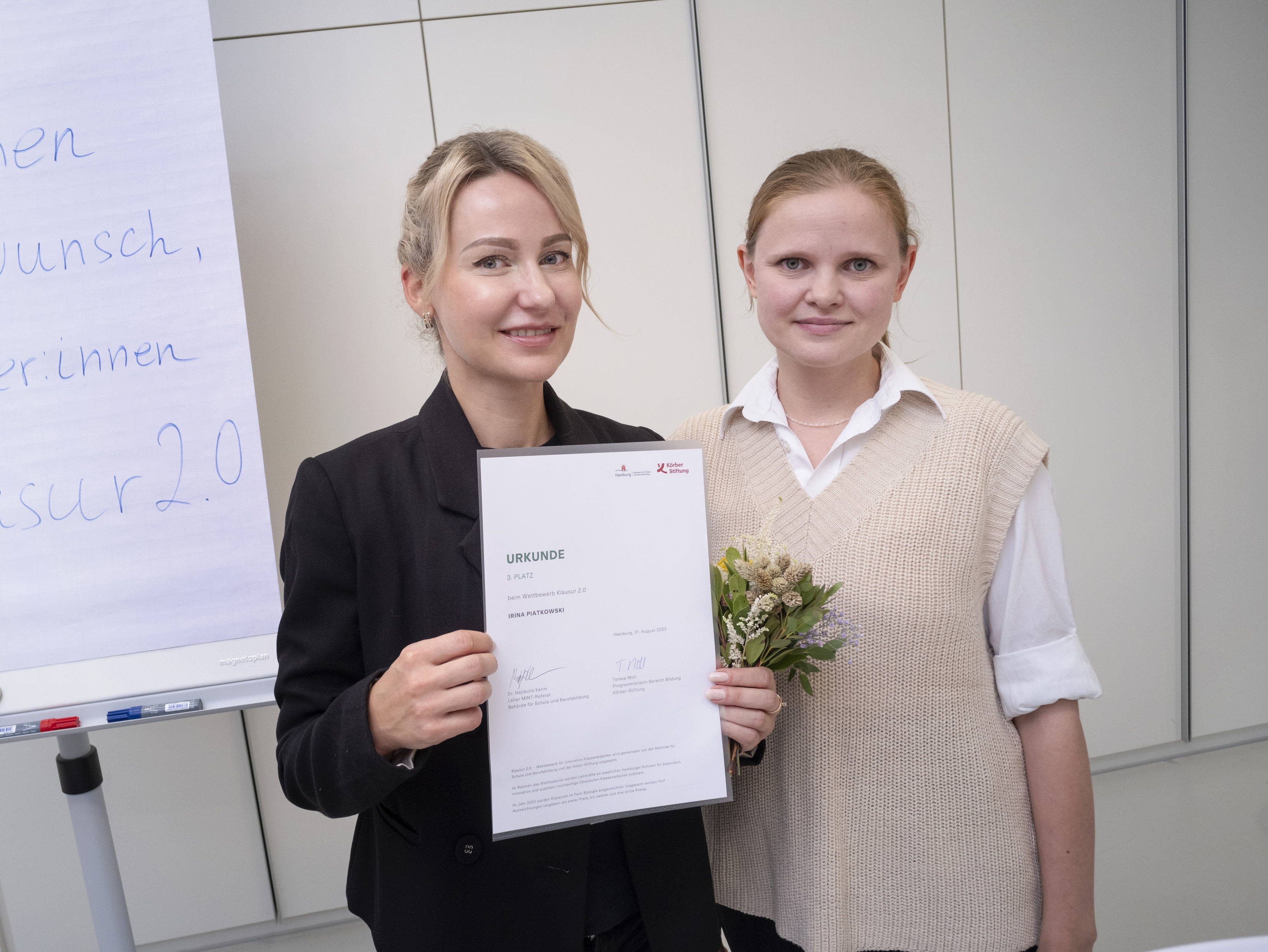 Zwei Frauen lächeln in die Kamera, eine hält eine Urkunde, die andere einen kleinen Blumenstrauß. Im Hintergrund steht ein Flipchart mit handgeschriebenem Text