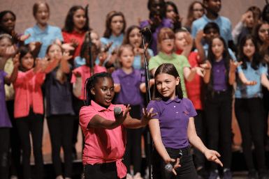 Ein Kinderchor mit bunten T-Shirts singt auf einer Bühne, während zwei Mädchen vorne mit Mikrofonen performen